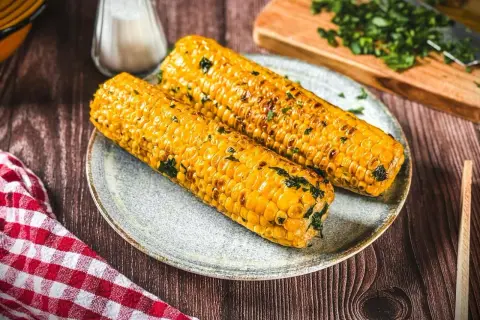 Zwei goldgelbe Maiskolben, perfekt gegrillt und mit Kräutern bestreut, liegen auf einem Teller. Ein Genuss für alle, die Maiskolben grillen lieben!