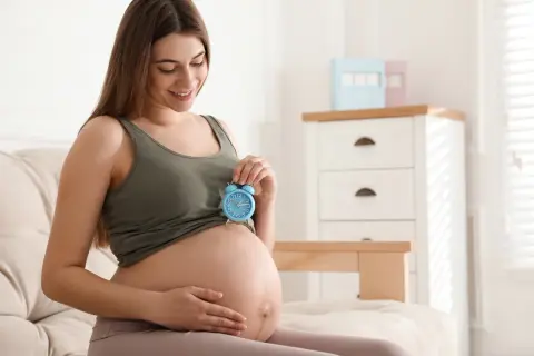 Uśmiechnięta kobieta w 9 miesiącu ciąży trzyma niebieski budzik na brzuchu. Czas na poród!