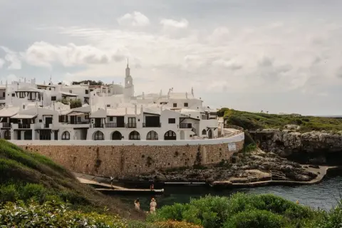Malownicze białe domki na Minorce, położone nad turkusową wodą, otoczone zielenią i skalistym wybrzeżem.