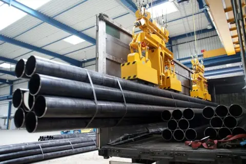 Żółte suwnice podnoszą stalowe rury w hali produkcyjnej. Budowa suwnicy jest kluczowa dla sprawnego transportu materiałów.