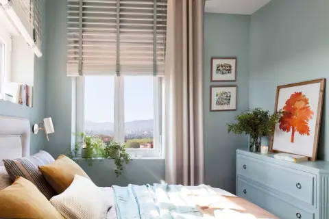 Acogedor plano dormitorio 9 metros cuadrados con cama, cómoda azul, cuadros y vistas a la ciudad.