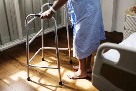 Osoba w szpitalnej koszuli, wspierając się na chodziku, stawia pierwsze kroki po udarze.