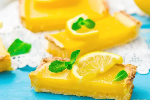 Fette di torta fredda al limone, guarnite con fettine di limone e menta fresca, su un tagliere blu.