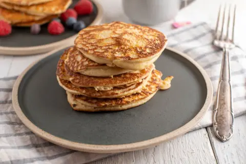 Stos puszystych fit pancakes z odżywki białkowej, ozdobiony malinami i borówkami, czeka na widelcu.