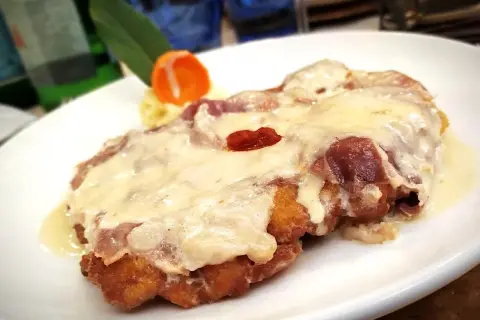 Cotoletta alla bolognese dorata e filante, con prosciutto e formaggio fuso, servita su un piatto bianco.