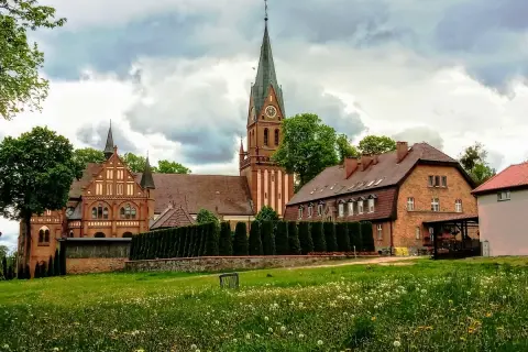 Majestatyczne sanktuarium w Gietrzwałdzie, z ceglaną architekturą i strzelistą wieżą, góruje nad zieloną łąką pełną kwiatów.