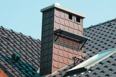 Czapka na komin dymowy chroni przed deszczem i ptakami. Widać ją na dachu z brązowej dachówki.