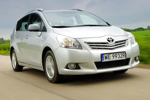 Srebrna Toyota Verso jedzie po drodze, otoczona zielonymi polami.