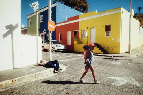 Une jeune fille porte son petit frère dans cette photo de rue colorée. Un homme dort sur le trottoir.