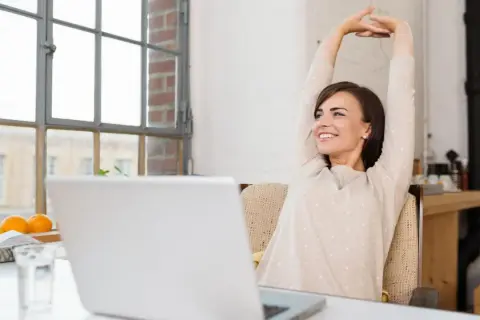 Frau streckt sich glücklich vor ihrem Laptop. Endlich Zeit für die 45-minütige Pause.