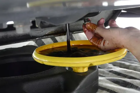 Mano sporca d'olio che tiene il tappo di scarico mentre l'olio scuro cola in una bacinella gialla. Forse c'era troppo olio nel motore.