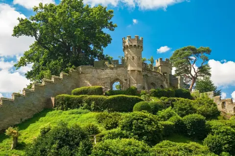 Warwick Castle reser sig stolt på en grönskande kulle, omgiven av frodig grönska och en klarblå himmel med vita moln.