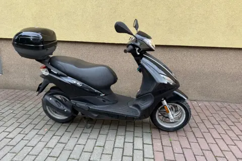 Czarny skuter Piaggio Fly 50 z kuferkiem na bagaż, zaparkowany na chodniku z kostki brukowej.
