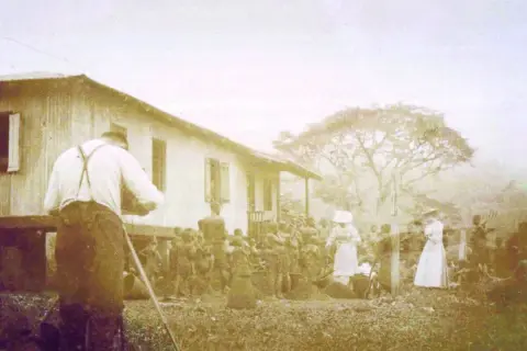 Une photo historique choquante : un homme filme une scène où des femmes en blanc semblent diriger un groupe d'indigènes devant une maison.
