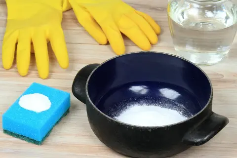 Gants jaunes, éponge bleue avec bicarbonate, casserole noire et un bocal de liquide. Astuces pour savoir comment nettoyer une casserole qui a brûlé.