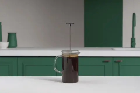 En kaffepress med mörkt kaffe redo att pressas. Köksbänk med gröna skåp och en kran i bakgrunden.