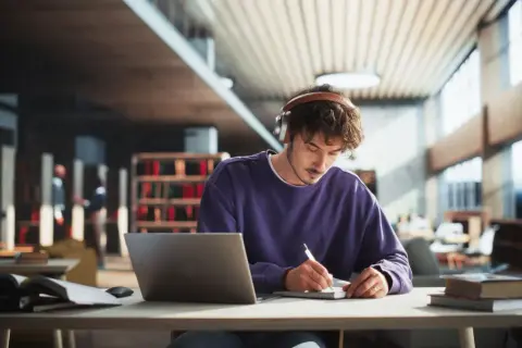 Junger Mann mit Kopfhörern lernt konzentriert in einer Bibliothek. Sein **online Studium Bayern** läuft gut.