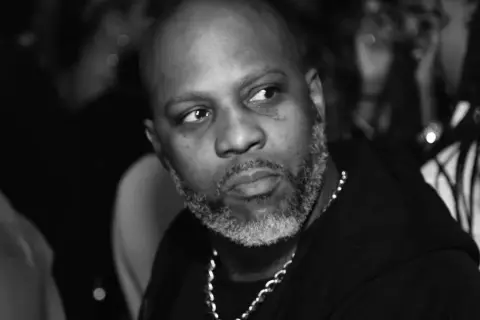 DMX, z siwą brodą i łańcuchem na szyi, spogląda w bok. W tle widać niewyraźne sylwetki, być może jego dzieci.