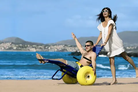 Coppia felice sulla spiaggia, lui su una sedia job con ruote gialle, lei lo spinge. Mare e collina sullo sfondo.