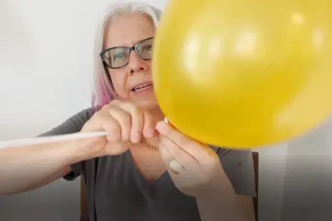 Kobieta w okularach mocuje żółty balon foliowy do białego patyka.