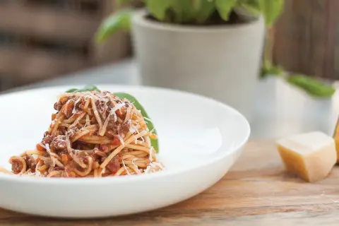 Pyszne spaghetti bolognese przepis oryginalny, podane na białym talerzu, posypane parmezanem. Obok kawałek sera i bazylia.