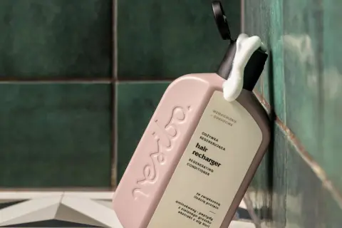 Omo jakie odżywki! Różowa butelka regenerującej odżywki do włosów "hair recharger" z pianką wydobywającą się z otwartego dozownika, na tle zielonych, łazienkowych płytek.