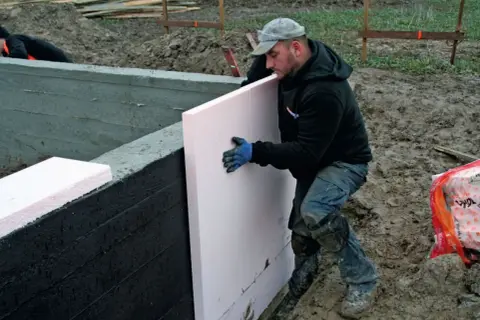 Budowlaniec układa styropian na ścianie fundamentu. To kluczowy etap, jak położyć fundament, zapewniający izolację.