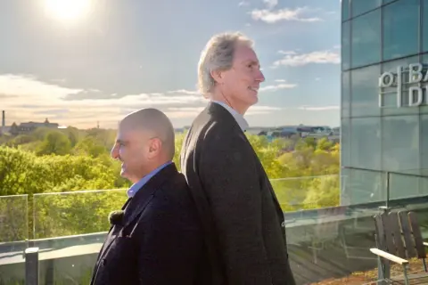 Erik Selin och en kollega står rygg mot rygg på en takterrass med utsikt över en grönskande stad.