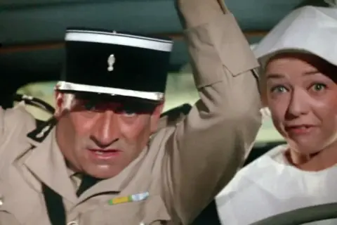 Louis de Funès w filmach, w mundurze żandarma, z przerażoną miną obok kobiety w białym nakryciu głowy.
