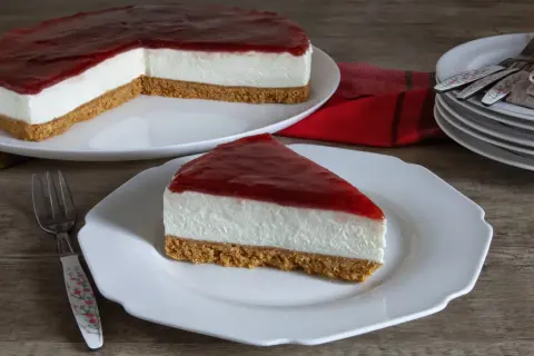 Una fetta di cheesecake fredda con copertura rossa su un piatto bianco, accanto a una torta intera.