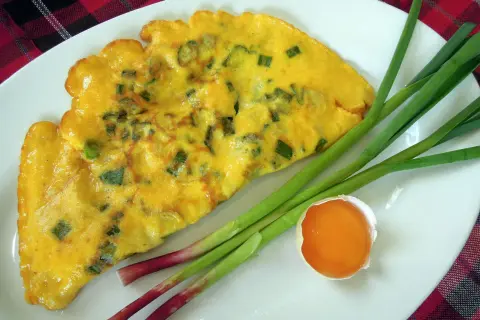 Une omelette à l'aillet, garnie de ciboule verte, est servie sur une assiette blanche avec des tiges d'aillet et un jaune d'œuf.