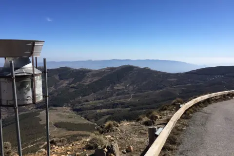 Vista panorámica desde la tetica de bacares, con un pluviómetro en primer plano y un paisaje montañoso y boscoso al fondo bajo un cielo azul.