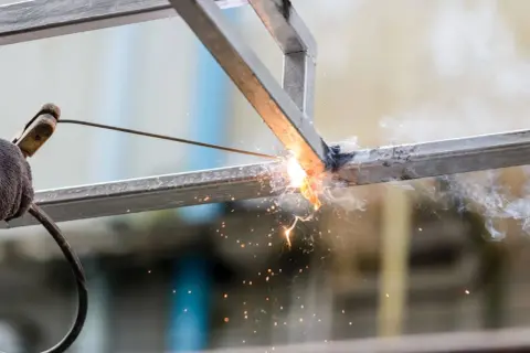 Spawanie profili metalowych, iskry i dym unoszą się w powietrzu.