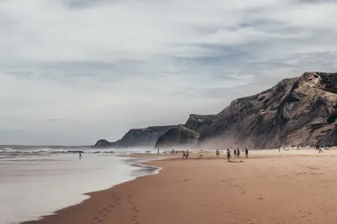 Plaża w Faro, wrzesień. Ludzie grają w piłkę, spacerują po piasku. W tle skaliste klify i ocean.