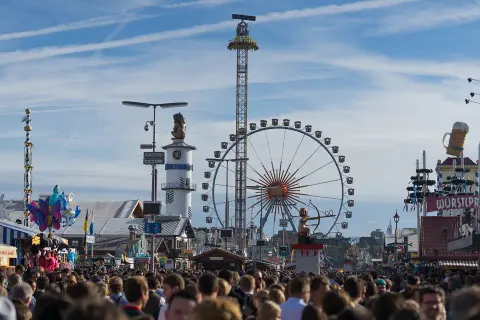 Menschenmassen auf dem **Bayern Oktoberfest** mit Riesenrad, Fahrgeschäften und Bierkrügen.