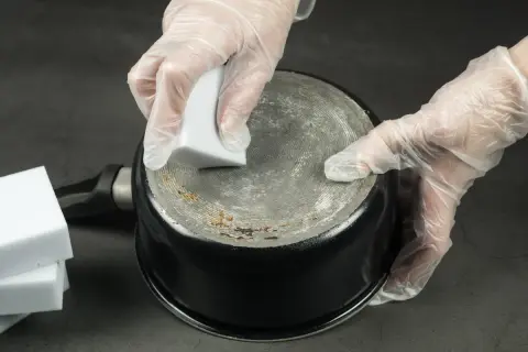 Ręce w rękawiczkach czyszczą przypalony garnek magiczną gąbką.