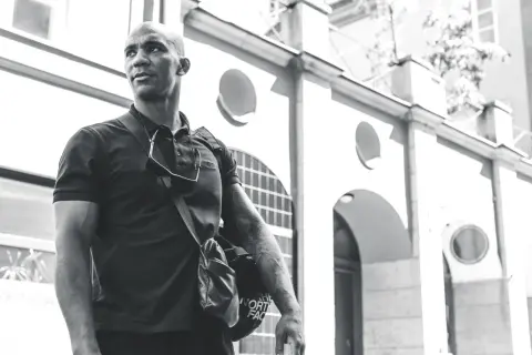 Nico Musoke, en man med tatueringar, bär en väska framför en byggnad i svartvitt.