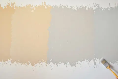 Échantillons de peinture sur un mur texturé, montrant 2 ou 3 couches de peinture dans des tons de beige et de gris. Un pinceau est visible à droite.