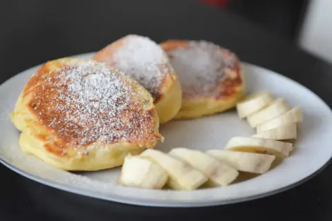 Szybkie placuszki na śniadanie, posypane cukrem pudrem, podane z plasterkami banana na białym talerzu.