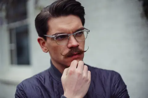 Un homme au visage losange, lunettes fines et moustache élégante, réfléchit.