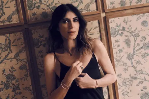 Woman in black slip dress leans against a wall with floral wallpaper.