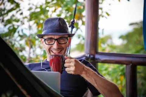 Mann mit Hut und Brille genießt Kaffee im Home Office English, mit Laptop und Kopfhörern.
