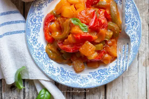Piatto di peperonata con patate, colorata e saporita, guarnita con basilico fresco su un piatto decorato.
