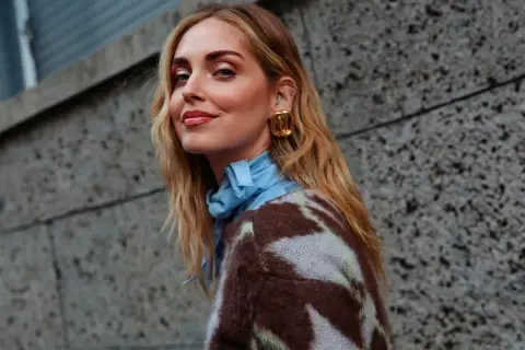 Mujer con media melena rubia, pendientes dorados y jersey estampado, sonriendo ante un muro de hormigón.