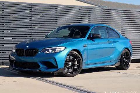 Błękitne BMW M2 Competition z czarnymi felgami i detalami, zaparkowane przed nowoczesną, szarą ścianą.
