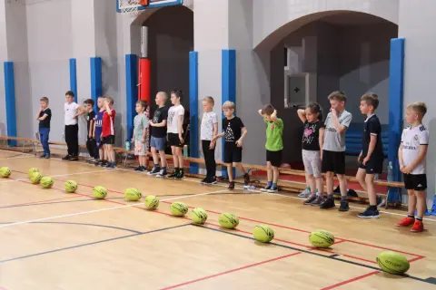 Dzieci stoją w rzędzie na sali gimnastycznej, gotowe do prostych konkurencji sportowych. Przed nimi leżą żółte piłki.