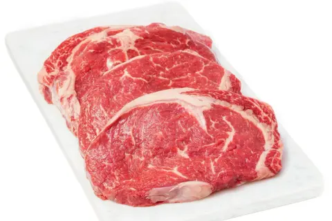 Tre råa, marmorerade entrecôte-biffar på en vit skärbräda, redo för medium rare temp.