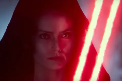Rey Skywalker w kapturze, z ponurym wyrazem twarzy, trzyma podwójny czerwony miecz świetlny.
