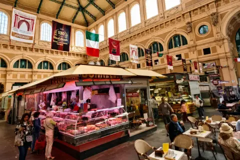 Interno di un vivace mercato delle vettovaglie, con banchi di carne, prodotti freschi e persone che fanno acquisti.