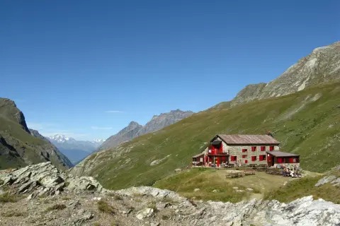 Rifugio nel Val di Rhemes, con persone sedute ai tavoli all'esterno, circondato da verdi pendii montuosi sotto un cielo azzurro.